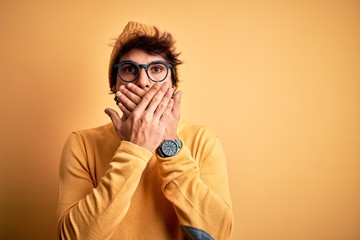 Young handsome man wearing casual t-shirt and glasses over isolated yellow background shocked covering mouth with hands for mistake. Secret concept.