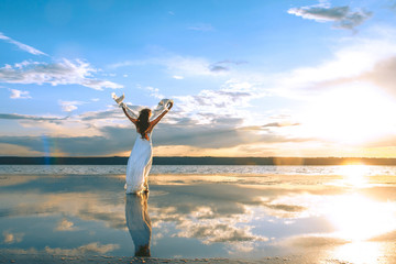 Fototapeta na wymiar girl in a white dress stands in the water at sunset holding hands up