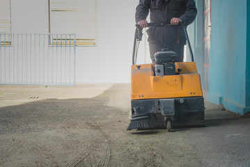 A person is pushing an orange gasoline powered industrial sweeper over dirty asphalt ground in an...