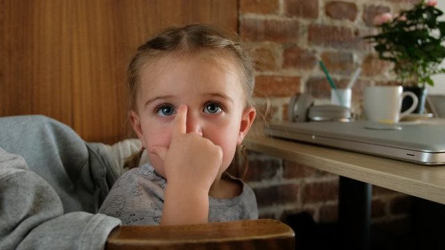 Cute little girl touches nose while sitting on chair near desk with laptop at home