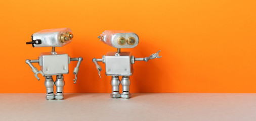 Robot points direction to second robot. Two simplified metal silver robotics toys on orange wall, gray floor background. Copy space