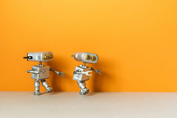 Two metal silver robots are walking. Simplified symbolic toy robotic characters on an orange...