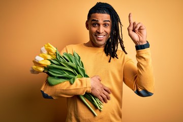 Young african american afro romantic man with dreadlocks holding bouquet of yellow tulips smiling amazed and surprised and pointing up with fingers and raised arms.