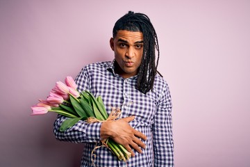 Young african american afro romantic man with dreadlocks holding bouquet of pink tulips skeptic and...