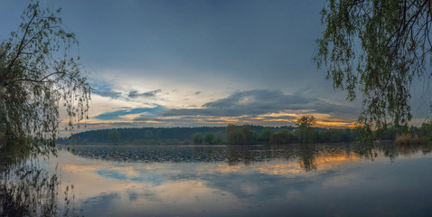 landscape panorama of a large beautiful lake and forest against a sunset with clouds with two large willows. Pond 14, known as the 5 glade. Kyiv, Ukraine