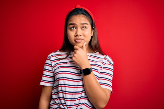 Young beautiful asian girl wearing casual striped t-shirt over isolated red background Thinking worried about a question, concerned and nervous with hand on chin