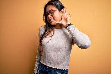 Young beautiful asian girl wearing casual sweater and glasses over yellow background smiling with hand over ear listening an hearing to rumor or gossip. Deafness concept.
