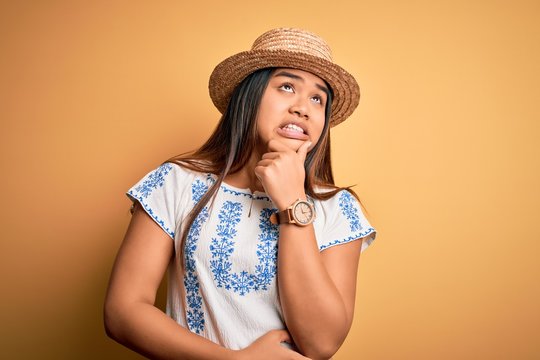 Young beautiful asian girl wearing casual t-shirt and hat standing over yellow background Thinking worried about a question, concerned and nervous with hand on chin