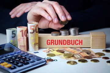 Grundbuch. Männliche Hand stapelt Geld-Turm (Euro). Begriff an Baustein. Münzen, Scheine &...