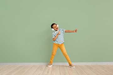 Little African-American boy listening to music and dancing against color wall