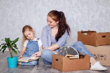 Fototapeta na wymiar Mother and daughter sort things out after moving to a new house or apartment. A woman and a little girl sit on the floor among cardboard boxes and read a book.