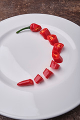 Red, searing, chilli pepper cut into pieces lies on a white plate on a brown background.
