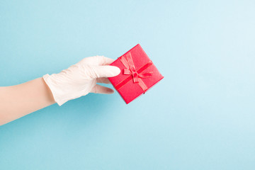 a hand in a blue disposable glove holds a red gift box with a bow from a red ribbon with gold thread, light blue background, copy space