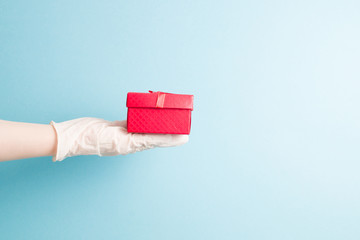 a hand in a blue disposable glove holds a red gift box with a bow from a red ribbon with gold thread, light blue background, copy space