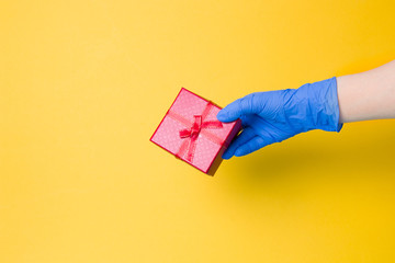 a hand in a blue disposable glove holds a red gift box with a bow from a red ribbon with gold thread, yellow background, copy space