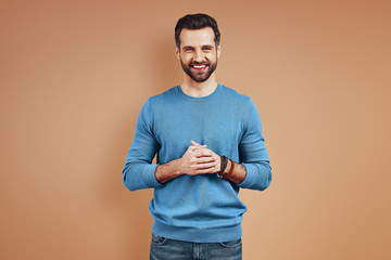 Charming young man in casual wear looking at camera and smiling while standing against brown background