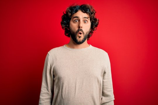 Young handsome man with beard wearing casual sweater standing over red background afraid and shocked with surprise expression, fear and excited face.
