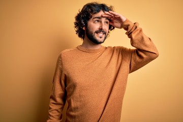 Young handsome man with beard wearing casual sweater standing over yellow background very happy and smiling looking far away with hand over head. Searching concept.