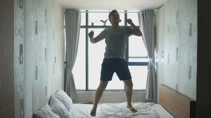 Handsome joyful man have fun, jumping, dancing on bed by the window
