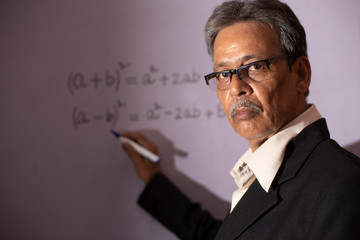 An senior man with formal suit and glasses writing on white board while taking online classes during lock down period. Indian lifestyle and teaching in home quarantine.