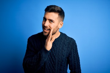 Young handsome man wearing casual sweater standing over isolated blue background hand on mouth...