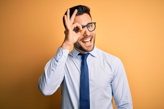 Young handsome businessman wearing tie and glasses standing over yellow background doing ok gesture with hand smiling, eye looking through fingers with happy face.