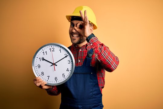 Young builder man wearing safety helmet holding big clock over yellow background with happy face smiling doing ok sign with hand on eye looking through fingers
