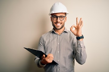 Young architect inspector man wearing builder safety helmet and using clipboard doing ok sign with...