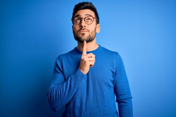 Young handsome man with beard wearing casual sweater and glasses over blue background Thinking concentrated about doubt with finger on chin and looking up wondering