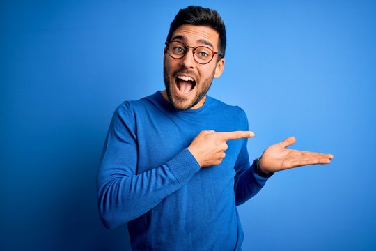 Young handsome man with beard wearing casual sweater and glasses over blue background amazed and smiling to the camera while presenting with hand and pointing with finger.