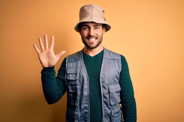 Handsome tourist man with beard on vacation wearing explorer hat over yellow background showing and pointing up with fingers number five while smiling confident and happy.