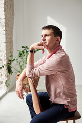 Young brunette man, wearing casual light pink jumper, sitting on brown stool in light room, thinking. Serious emotions.Three-quarter portrait of businessman, resting on his day off in loft apartment.
