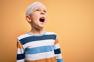 Young little caucasian kid injured wearing medical bandage on head over yellow background crazy and mad shouting and yelling with aggressive expression and arms raised. Frustration concept.