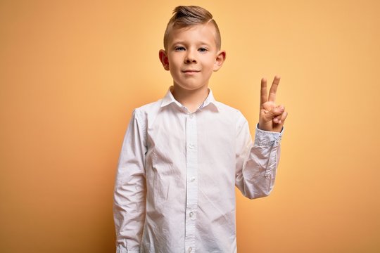 Young little caucasian kid with blue eyes wearing elegant white shirt over yellow background smiling with happy face winking at the camera doing victory sign. Number two.