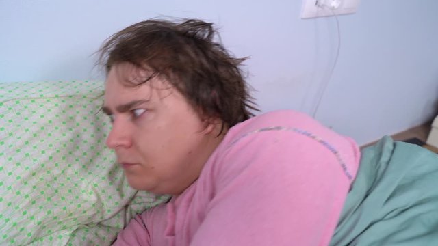 Shaggy untidy guy in pink pajamas wakes up suddenly and looks around with curiosity, he scaredly pushes someone nearby with hand. Young man has psychological problems causing nightmares.