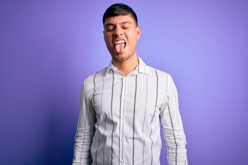 Young handsome hispanic man wearing elegant business shirt standing over purple background sticking tongue out happy with funny expression. Emotion concept.