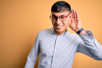 Young handsome hispanic business man wearing nerd glasses over yellow background smiling with hand over ear listening an hearing to rumor or gossip. Deafness concept.