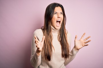 Young beautiful girl wearing casual turtleneck sweater standing over isolated pink background crazy and mad shouting and yelling with aggressive expression and arms raised. Frustration concept.