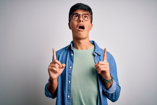 Young handsome man wearing casual shirt and glasses over isolated white background amazed and surprised looking up and pointing with fingers and raised arms.