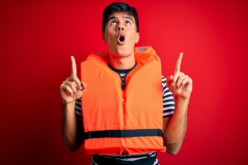 Young handsome man wearing orange safety life jacket over isolated red background amazed and surprised looking up and pointing with fingers and raised arms.
