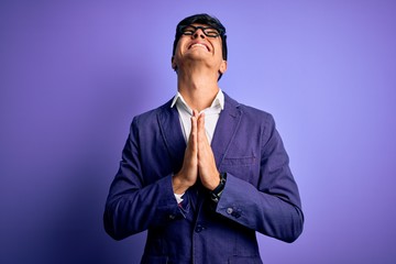 Young handsome business man wearing jacket and glasses over isolated purple background begging and...