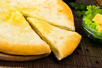 Ossetian pie with cheese on a wooden stand and a brown Mat, with ingredients. One piece was cut off.