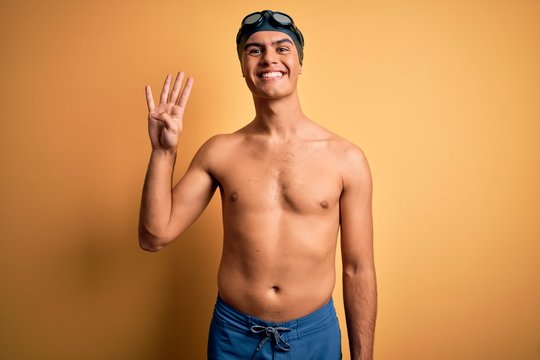 Young handsome man shirtless wearing swimsuit and swim cap over isolated yellow background showing and pointing up with fingers number four while smiling confident and happy.
