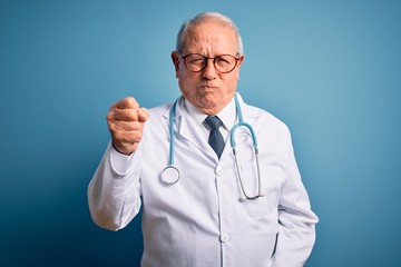 Senior grey haired doctor man wearing stethoscope and medical coat over blue background angry and...