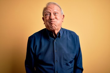 Grey haired senior man wearing casual blue shirt standing over yellow background puffing cheeks with funny face. Mouth inflated with air, crazy expression.
