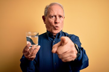 Grey haired senior man drinking a fresh glass of water over yellow isolated background pointing...
