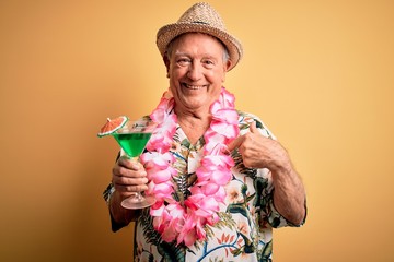 Grey haired senior man wearing summer hat and hawaiian lei drinking a cocktail with surprise face...