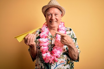 Grey haired senior man wearing summer hat and hawaiian lei holding paper plane on vacation very...