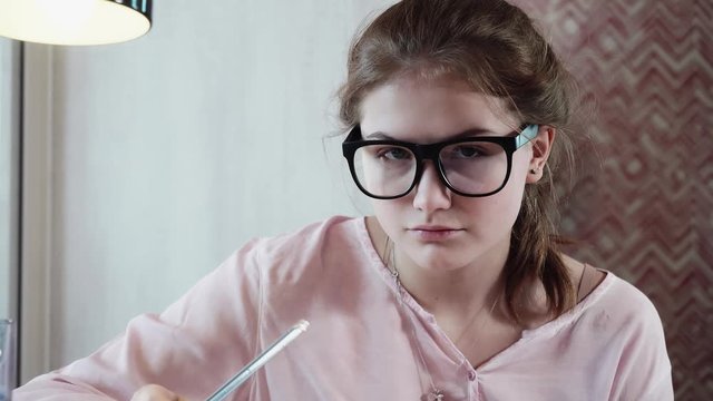Face of serious teeanger in glasses with black frame drawing picture sitting at table then she looks at camera and smiles. Close up view.