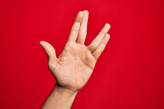 Hand of caucasian young man showing fingers over isolated red background greeting doing Vulcan salute, showing hand palm and fingers, freak culture